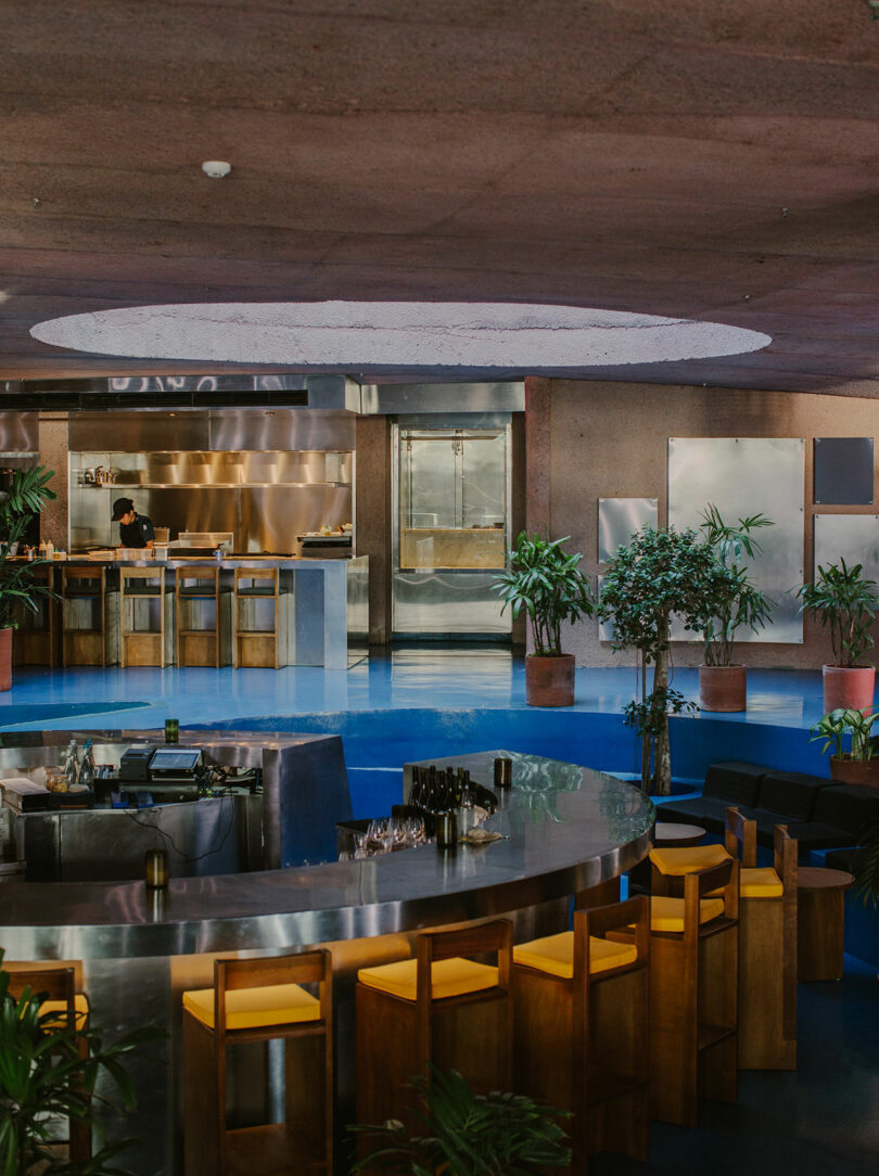 A modern bar with wooden stools and a circular counter is set in a spacious room, reminiscent of Desa Potato Head, with lush plants and a kitchen area in the background.