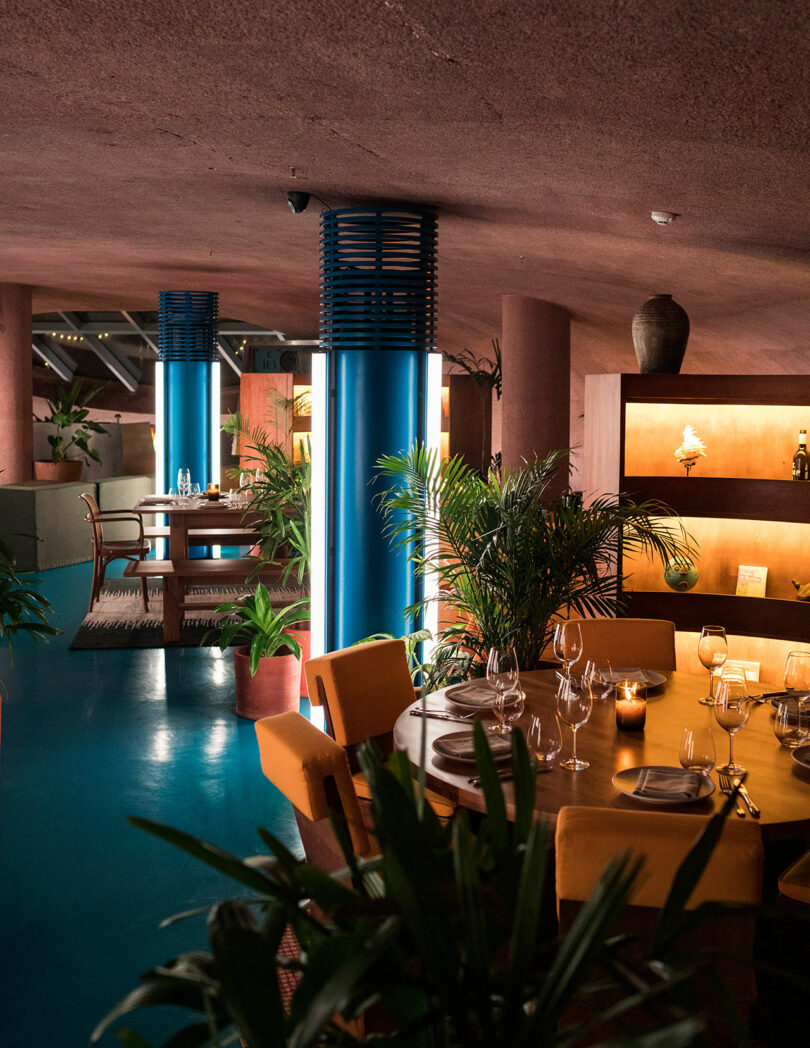 The modern interior of Desa Potato Head features a warm color scheme, with round tables set for dining, tall blue columns, and lush green plants.