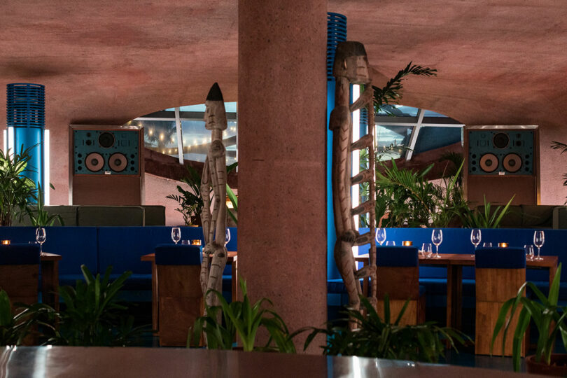 A dimly lit restaurant interior at Desa Potato Head features tribal statues, blue seating, wooden tables set with wine glasses, and lush greenery.