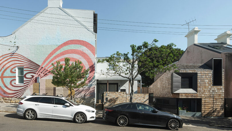 Two cars parked on a residential street; one white and one black. A house with a colorful mural is visible in the background alongside modern architecture and trees.