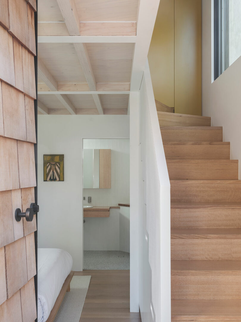 Interior showing a wooden staircase leading to an upper level, a bedroom with a white bed, and a partial view of a bathroom with a mirror and artwork on the wall.