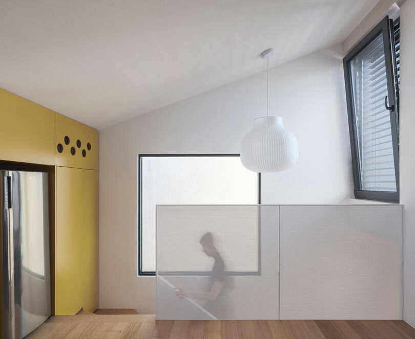 Modern interior with yellow cabinets, large stainless steel fridge, and a person blurred in motion behind a semi-transparent divider, near a window with a geometric pendant light overhead.