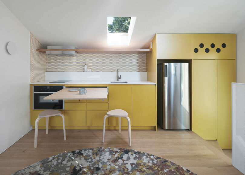 Modern kitchen with yellow cabinetry, a built-in oven, stainless steel fridge, and two wooden stools. A skylight provides natural light. Walls feature a white tile backsplash.