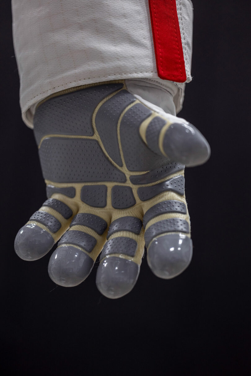 Close-up of a gloved hand in a spacesuit, with gray textured surfaces and padded fingertips against a dark background.