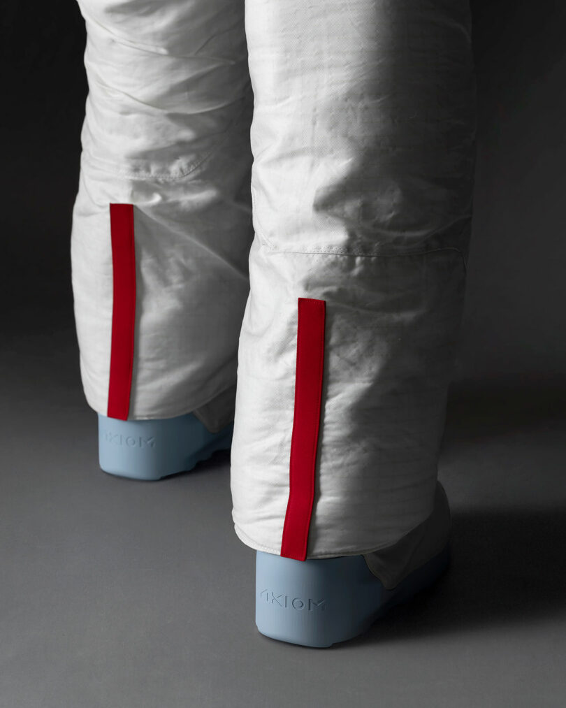 Close-up of white padded pants with red vertical stripes near the seams, worn over light blue boots labeled "AXIOM" at the heel.