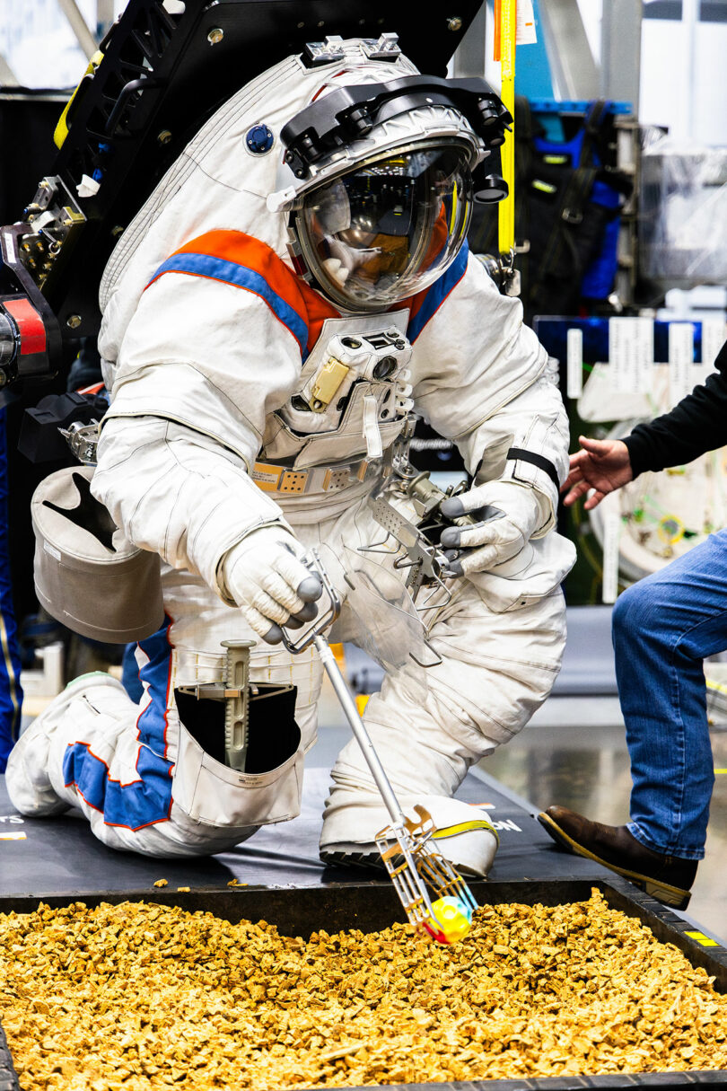 Astronaut in a training suit practices maneuvering with a tool in a simulated space environment indoors, interacting with objects on the ground.