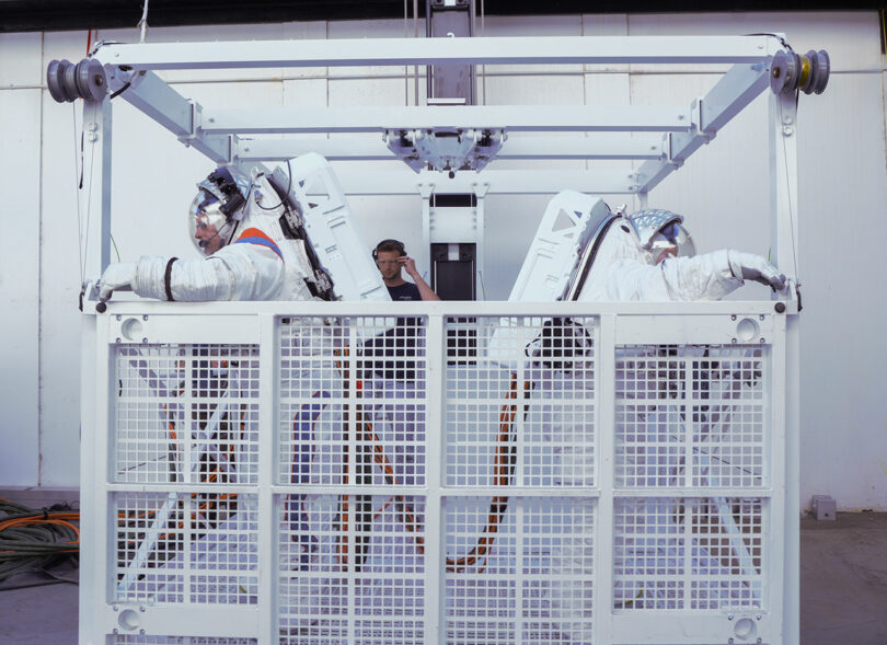 Two people in space suits are seated in a metal cage structure with a technician observing them from the background.