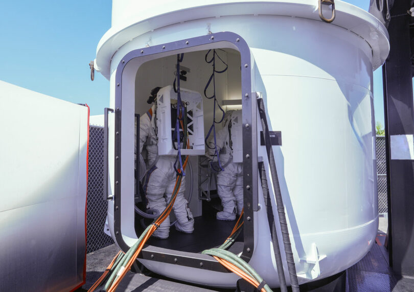 Two astronauts in spacesuits stand inside a white spacecraft module, with cables extending from the open doorway.