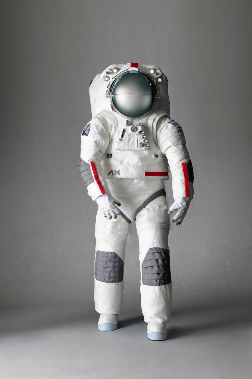 An astronaut suit displayed upright against a plain gray background. The suit features white and gray panels with red accents and a reflective helmet visor.