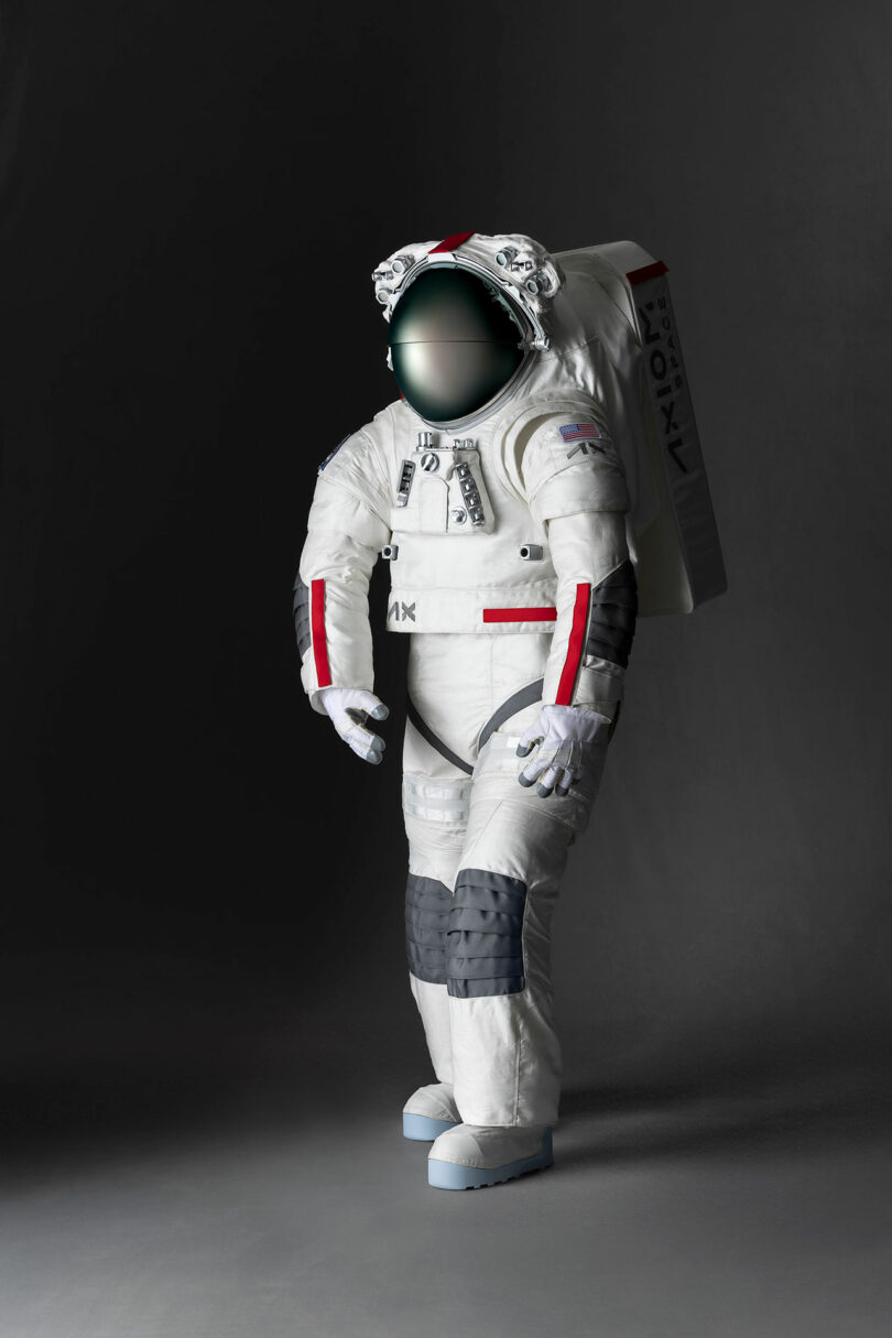 A person in a white and black advanced spacesuit with a large helmet visor stands in front of a dark background. The suit is marked with various technical details and logos.