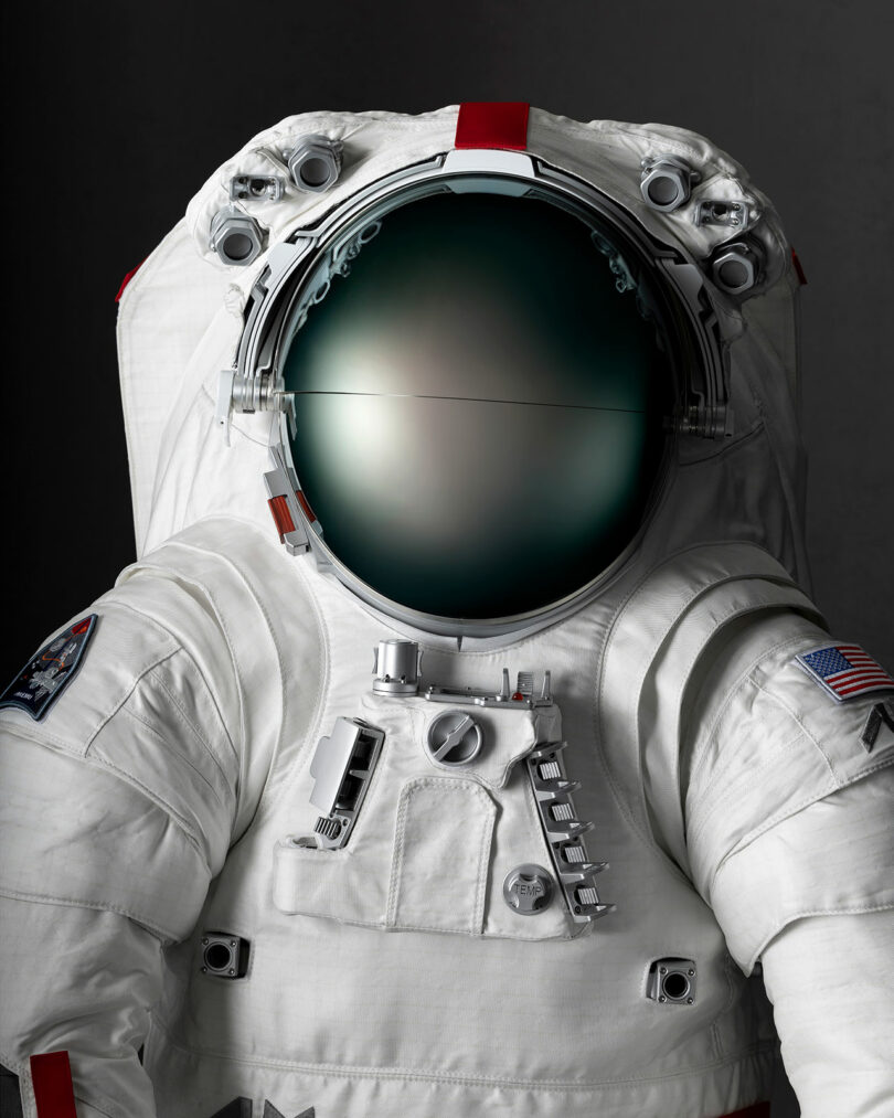 A detailed close-up of a spacesuit featuring the American flag, a reflective helmet visor, and various suit attachments against a dark background.