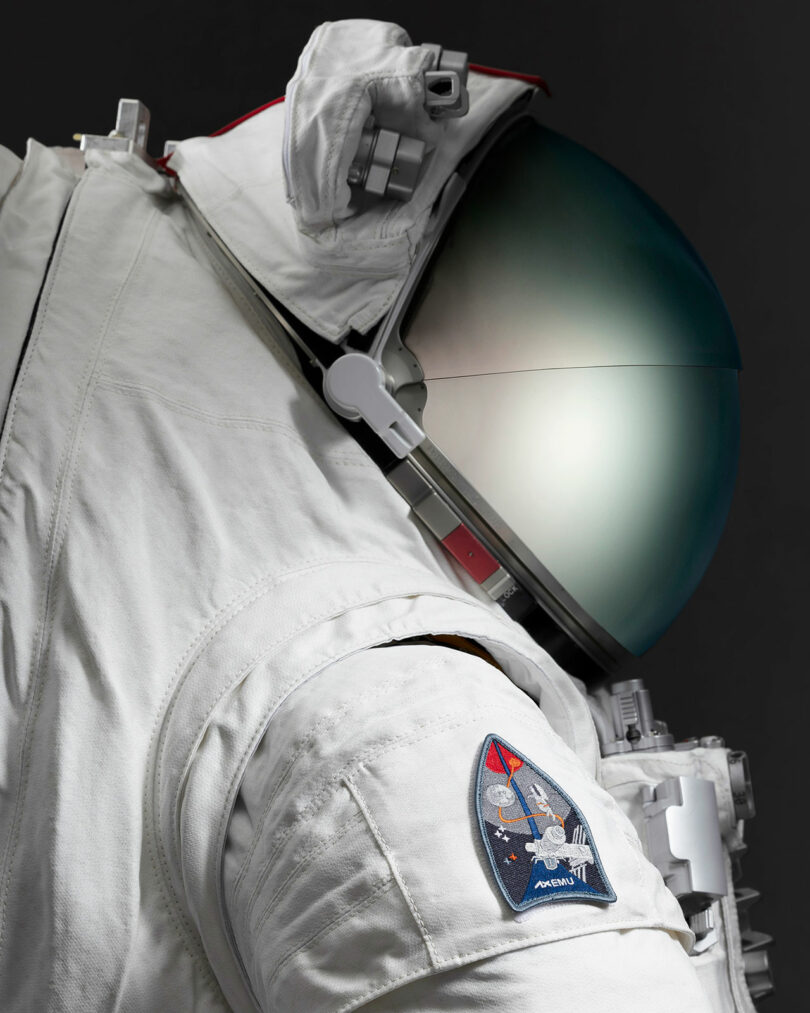 Close-up of an astronaut suit with a reflective helmet. The suit features a patch with a space-themed design on the shoulder.