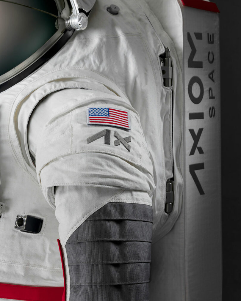 Close-up of a white spacesuit with an American flag patch and the "Axiom Space" logo. The suit features detailed stitching and joint padding.