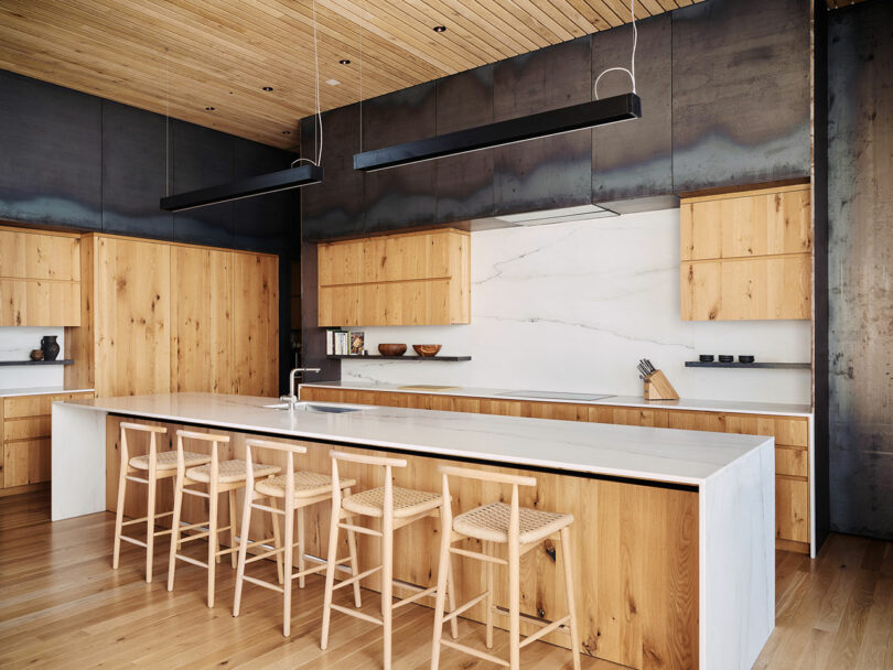 Modern kitchen with wooden cabinets, a large white marble island, and matching wooden bar stools. Ceiling and walls feature dark metallic elements, adding a contemporary design touch.