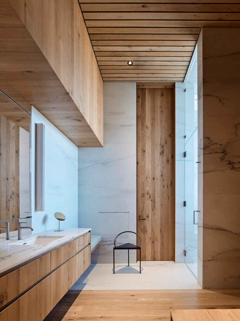 Modern bathroom with wood paneling, marble walls, large mirror, and glass shower enclosure. A small round chair sits in the middle of the room.