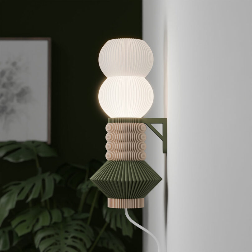 A modern STAK Modular Lamp with a layered, ribbed design and soft white illumination. The background highlights a dark green wall, a leafy plant, and framed artwork.