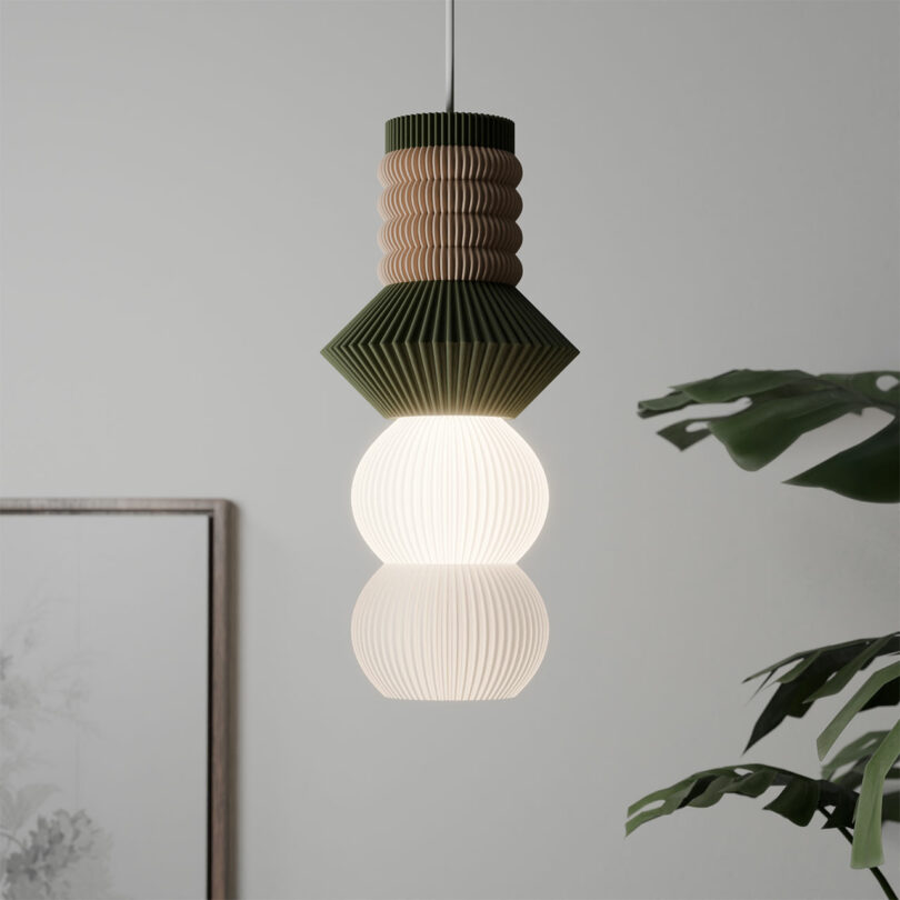 The STAK Modular Lamp, with its geometric, layered design, elegantly hangs in front of a framed picture and a plant.