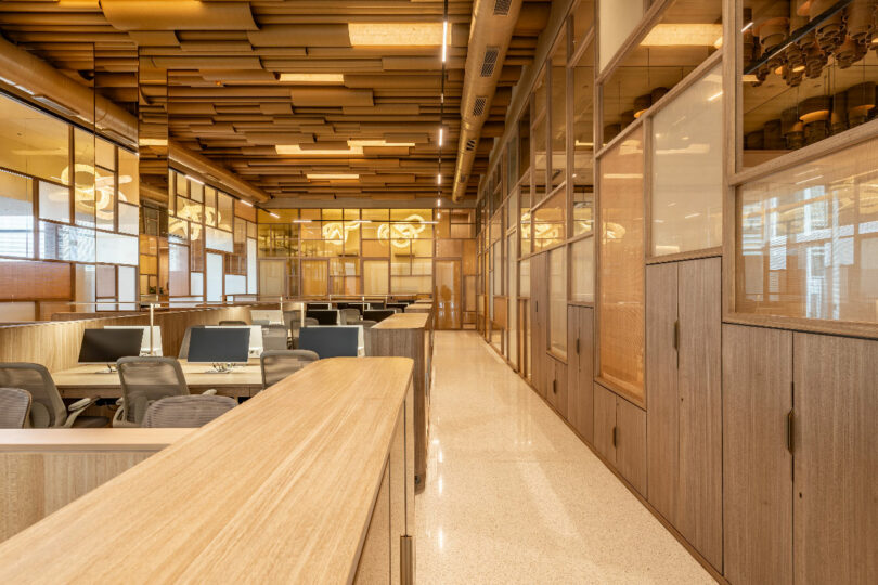 A modern office space with wooden furnishings, computer workstations, and a corridor leading to private rooms with glass and wood partitions.