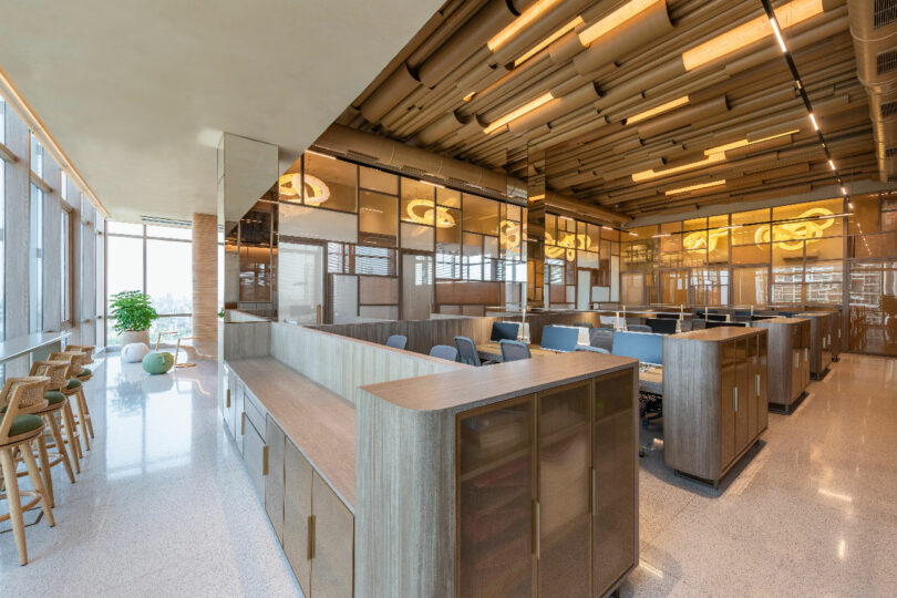 Modern open office space with wooden partitions, ergonomic chairs, workstations, overhead lighting, and large windows providing natural light. Bar stools and small tables are at the far left side.