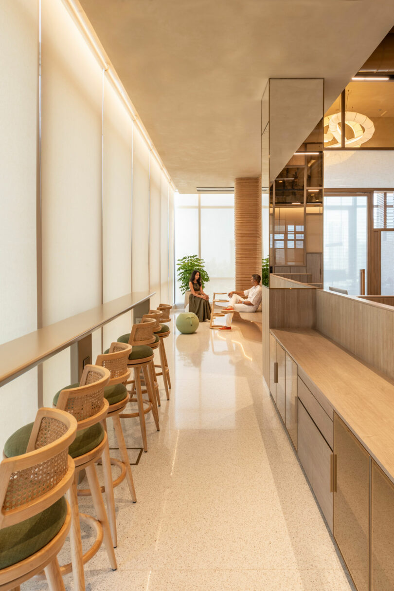 Modern lounge area with a long counter and bar stools facing floor-to-ceiling windows. In the background, two people are seated on a couch with a green gym ball nearby.