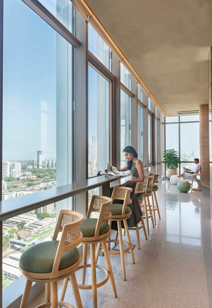 A person sits at a high counter reading a newspaper in a bright room with large windows offering a city view. Empty stools are lined up along the counter. Another person is in the background by a large plant.