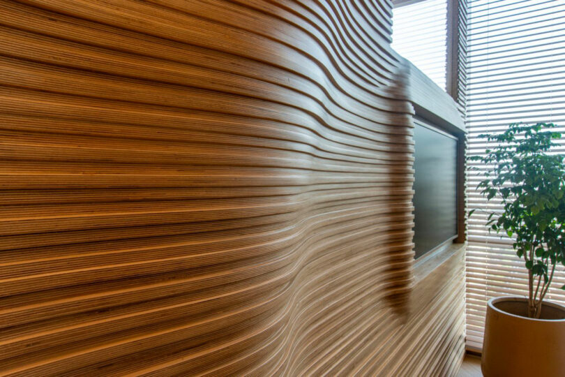 A wooden wall with wavy grooves and a built-in TV screen next to a potted plant by the window with blinds.