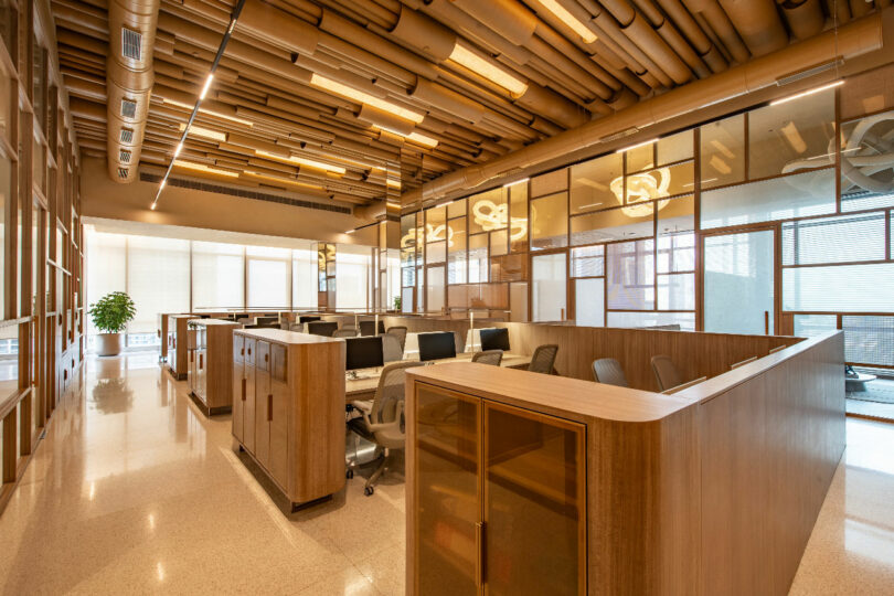 A modern, open-plan office space with wooden cubicles, ceiling ducts, and large windows letting in natural light. Empty desks are equipped with computers and chairs. A plant is in the background.