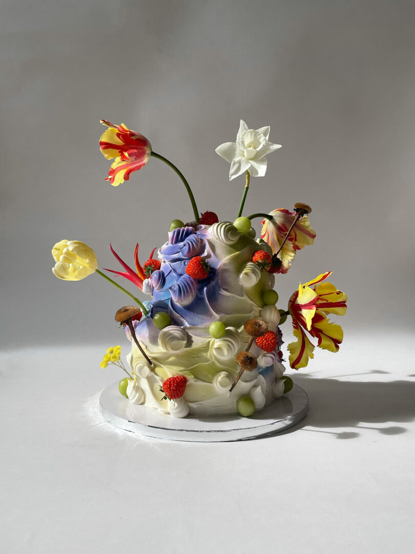 A colorful cake decorated with various flowers, strawberries, and green spheres on a white background.