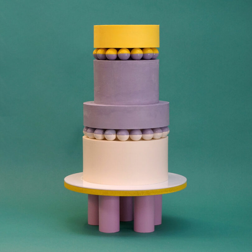 A four-tiered cake with cylindrical and spherical shapes in yellow, lavender, and cream colors, set against a green background.