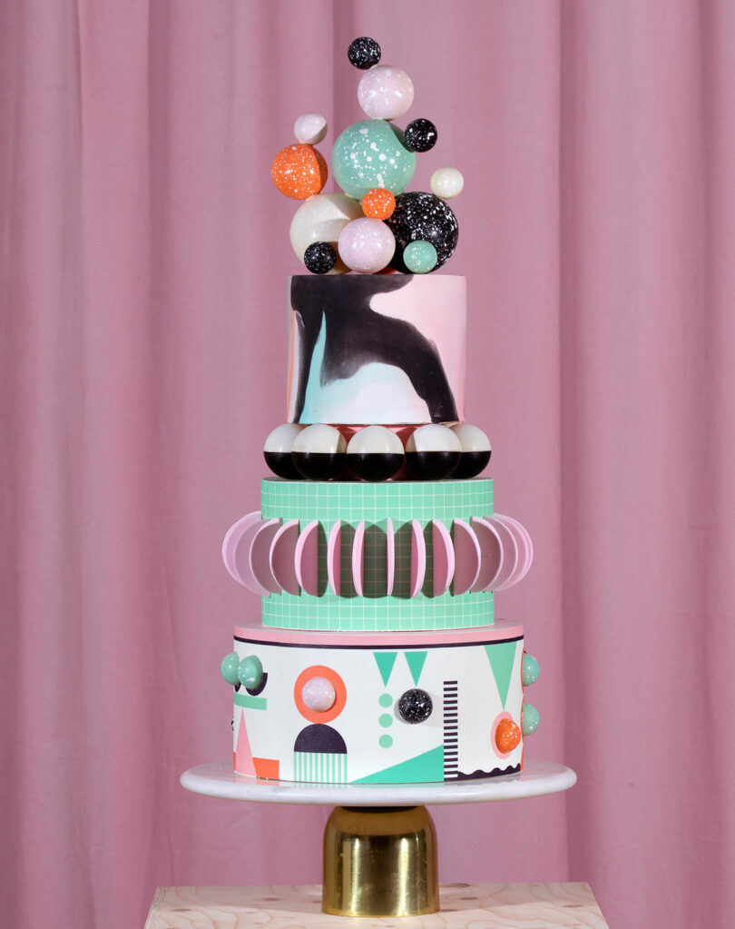 A four-tiered cake with geometric designs and a topper of colorful spheres, set against a pink curtain backdrop.