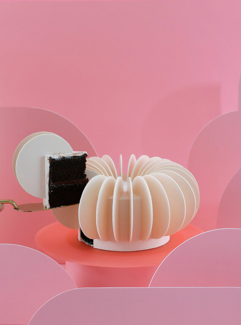 A slice is being cut from a round, layered chocolate cake with white frosting against a pink background.