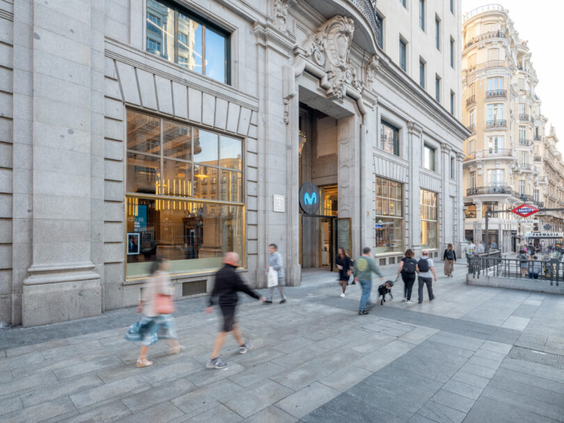 People walking on a busy city street past a large building with glass windows. A metro entrance with a blue "M" sign is visible on the right.