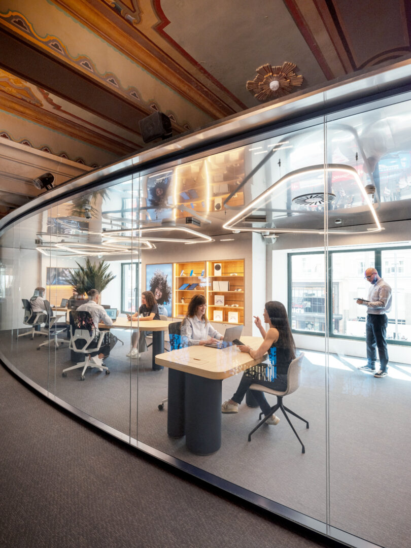 A modern office with glass walls, featuring people working at desks and one person standing by the window, set against an ornate ceiling.