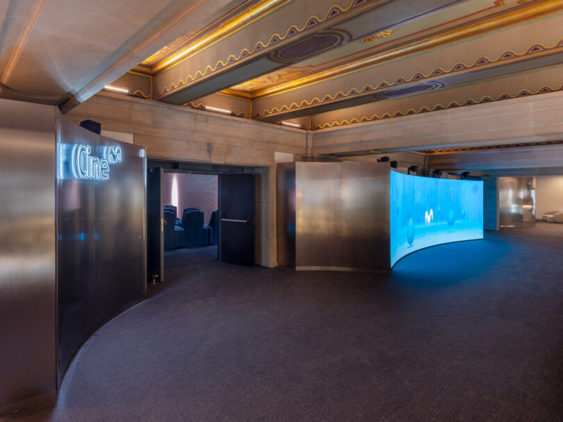 A modern cinema entrance with curved wall displays and ceiling lights, leading to a dimly lit theater room.