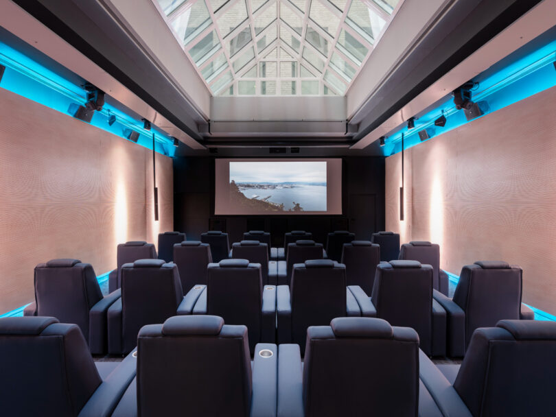 A modern home theater with black recliner seats, a large screen displaying a coastal scene, and a skylight ceiling.