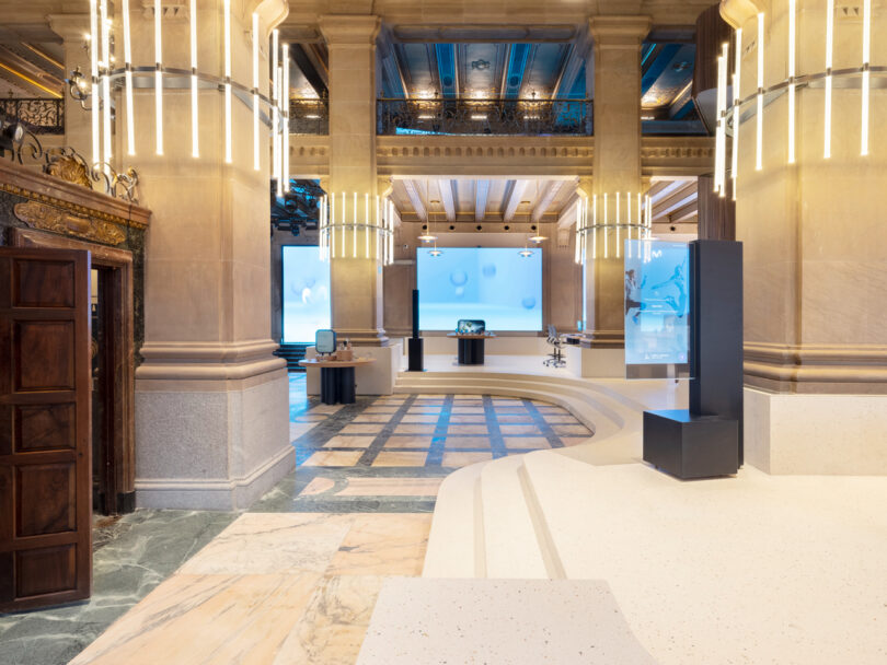 Grand, ornate building interior with high ceilings and large, cylindrical chandeliers. The space includes modern displays, a marble floor, and an intricate architectural design.