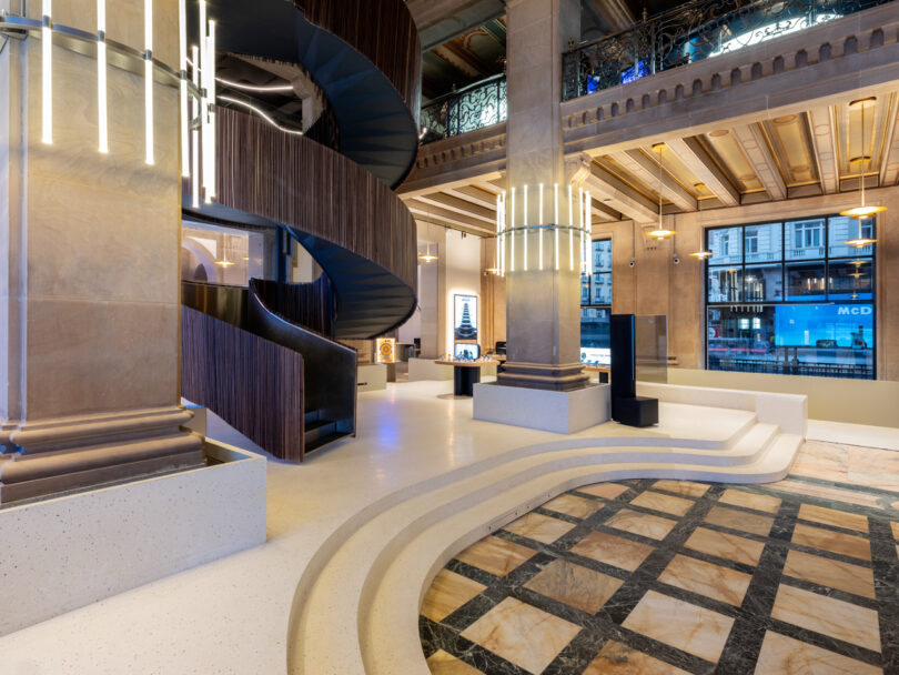 Spiral staircase in a modern building lobby with marble floors, high ceilings, and large windows displaying street views outside.