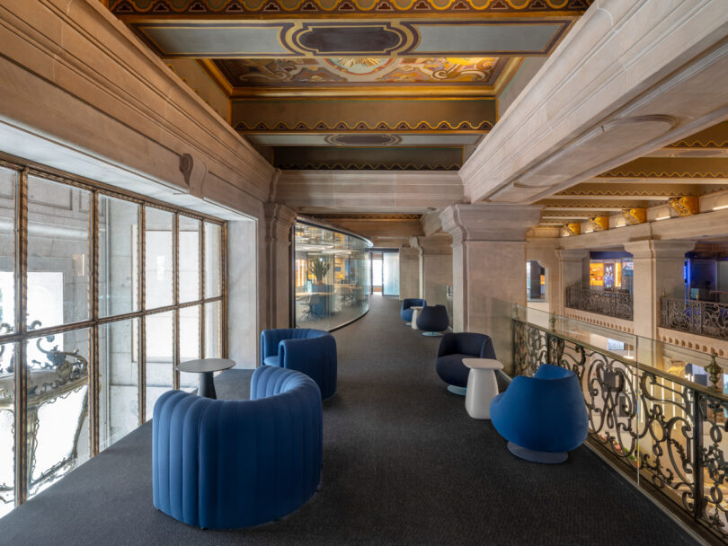 A hallway with cushioned seating, side tables, and an ornate ceiling. Large windows on the left and a glass-walled area ahead.