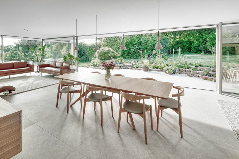 Modern dining room with a wooden table, eight chairs, pendant lights, and large windows overlooking a garden. The room is bright and minimalistic, featuring a lounge area with sofas.