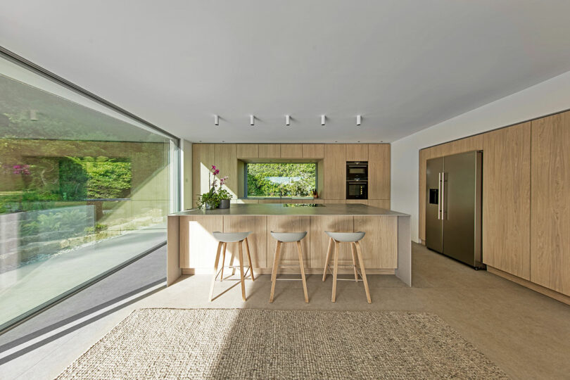 Modern kitchen with large glass window, wooden cabinetry, an island with three stools, stainless steel appliances, and a woven rug on the floor. Garden view outside.