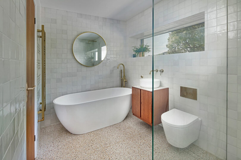 Modern bathroom with a freestanding bathtub, round mirror, wooden vanity with a sink, and a glass-enclosed shower. The walls are tiled, and there is a window overlooking greenery.