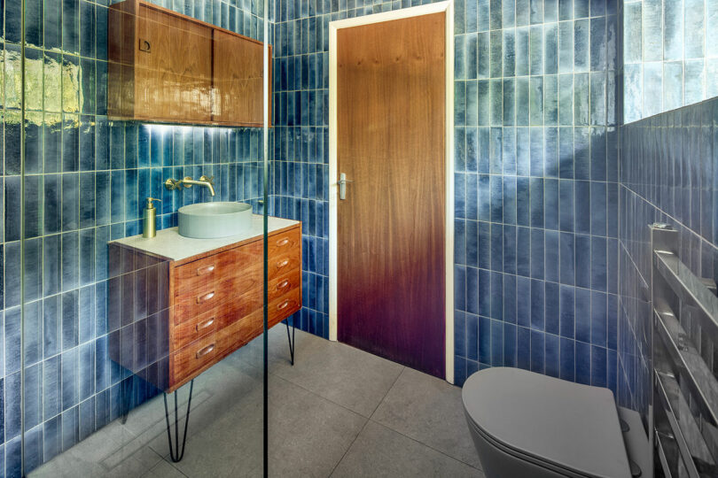 Bathroom with blue tiled walls, wooden door, wooden vanity, round sink, and a modern toilet.