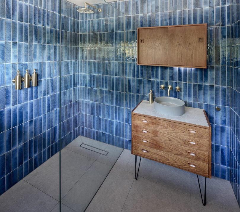 Modern bathroom with blue tiled walls, a wooden vanity with a round sink, wall-mounted faucets, and a walk-in shower with a rainfall showerhead.