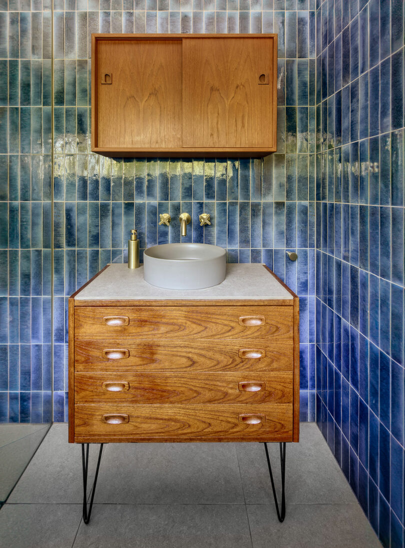 A bathroom with blue tiled walls features a wooden vanity with a round sink, wall-mounted faucet, and a wooden cabinet above.