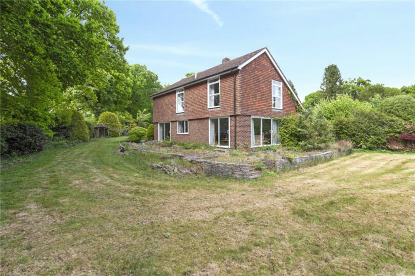 A two-story brick house with large windows, surrounded by overgrown grass and lush trees, under a clear blue sky.