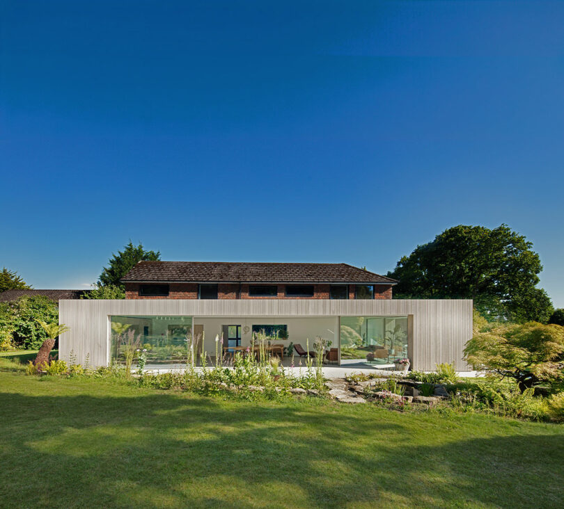 Modern glass-walled house with a garden in front, set against a brick building and a clear blue sky.