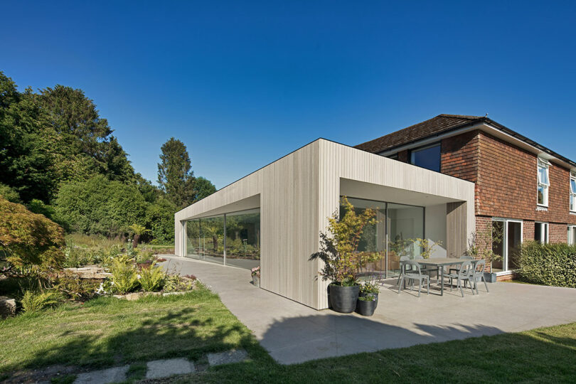 Modern house extension with large glass windows, surrounded by greenery. Outdoor seating area on the patio. Clear blue sky above.