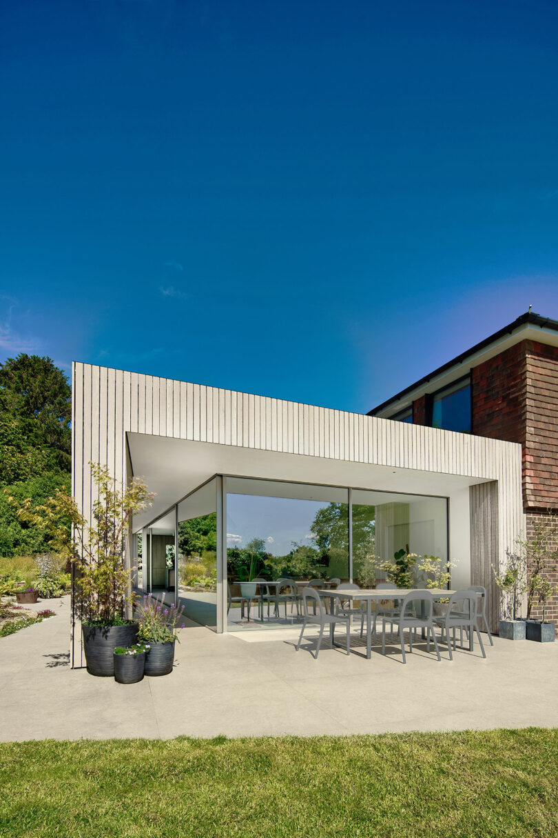 Modern patio with glass walls, outdoor dining table, and chairs. Surrounded by potted plants and greenery, under a clear blue sky.