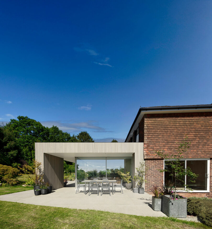 Modern house with a glass extension, featuring a dining table and chairs on a concrete patio, surrounded by plants, set against a clear blue sky and green foliage.