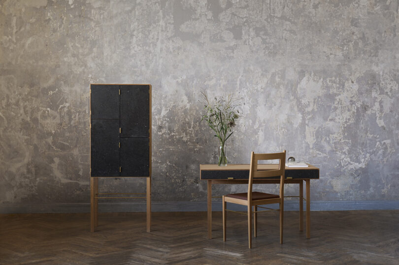 A minimalist room with a textured grey wall, featuring a wooden cabinet, a desk with a chair, and a vase with flowers. The floor is herringbone wood.